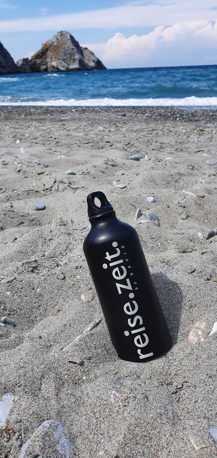 Eine schwarze Thermosflasche mit der Aufschrift reise.seit steckt im Sand und dahinter das Meer mit einem Felsen