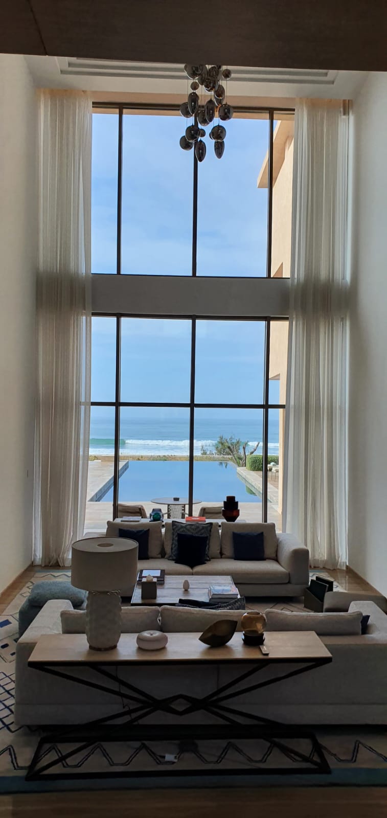 Ein luxuriöses Wohnzimmer mit Blick durch ein schwarz gerahmtes Sprossenfenster auf eine Terrasse mit Pool und dahinter das weite Meer