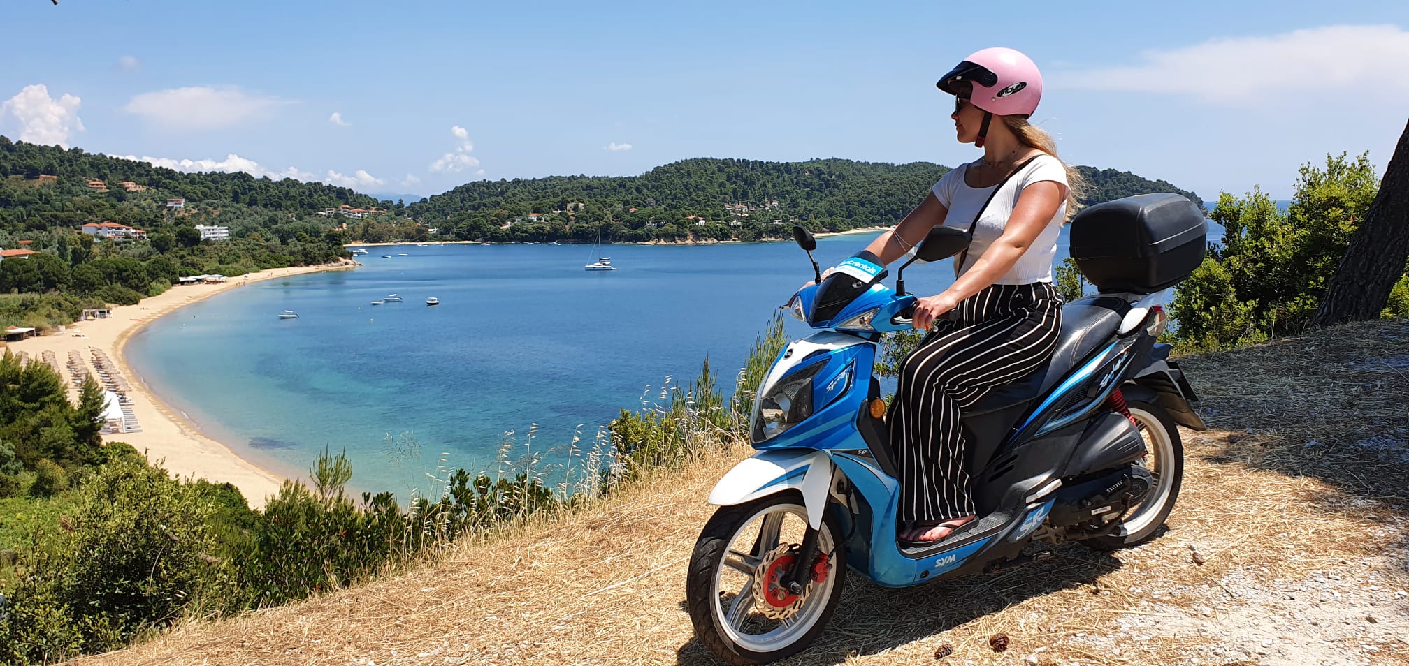 Eine sommerlich gekleidete Frau mit einem rosa Helm sietzt auf einem blauen Motorroller und schaut auf eine sonnenverwöhnte Bucht mit türkisblauem Meer hinab